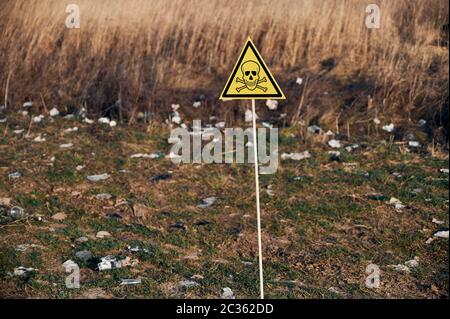 Yellow triangle with skull and crossbones symbol warning about poisonous substances and danger on abandoned territory with trash. Garbage waste field with toxic hazard sign Stock Photo