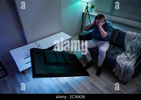 Man Sitting On Sofa In Front Fallen Television With Broken Screen Stock Photo