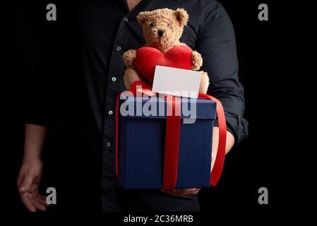 adult man in a black shirt holds a blue square box tied with a red ribbon and sits on top of a brown teddy bear with a heart, concept of congratulatio Stock Photo