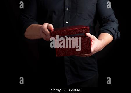 adult man in a black shirt holds a cardboard red gift box and opens the lid, dark background Stock Photo