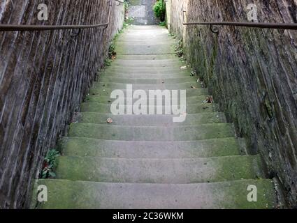 a perspective view of narrow old outdoor stone steps descending between walls with weeds and moss growing in the corners Stock Photo