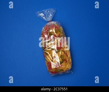 transparent cellophane bag with raw colorful pasta in the form of bows on a blue surface, copy space Stock Photo