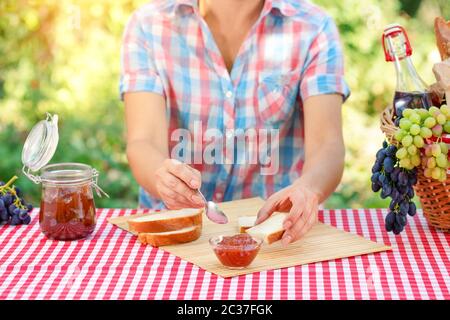 A woman in a plaid shirt spreads jam on bread. Red tablecloth, picnic basket, grapes. Sunny day. Picnic concept Stock Photo