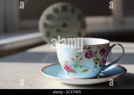 English blue and white tea cup on saucer in fine china with delicate flowers and gold edging. Clock in Background. Porcelain Stock Photo