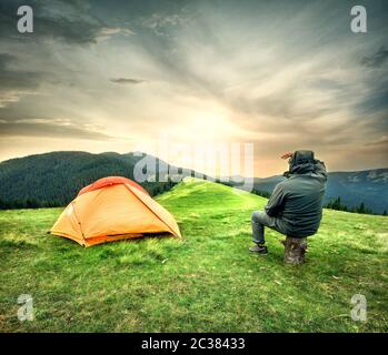 Man sitting near tent looking at the setting sun over mountains Stock Photo