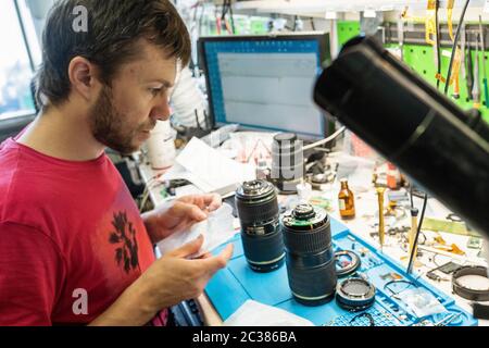 Master checks photo lenses in repair shop. Bearded man in red t-shirt works at workplace with master's tools on it. Service, repair and maintenance of Stock Photo