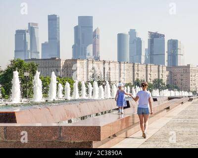 A little baby girl walks along the edge of the fountain on a sunny summer day by the hand with her mother against the backdrop of modern skyscrapers Stock Photo