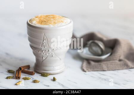A cup of chai latte with cane sugar on top. On a marble table with white background. There are some cardamom pods and cinnamon sticks next to the whit Stock Photo