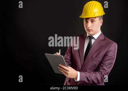Portrait of engineer architect builder with blond hair wears a yellow safety helmet dressed in burgundy jacket, white shirt and black tie takes notes Stock Photo
