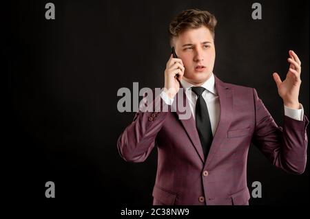 Law student with blond hair dressed in burgundy jacket, white shirt and black tie displeased face and gestures and talking on cellphone posing on isol Stock Photo