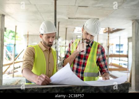 Building site. Building workers in yellow vests brainstorming over blueprint, discussing it, one of men thouching his chin Stock Photo