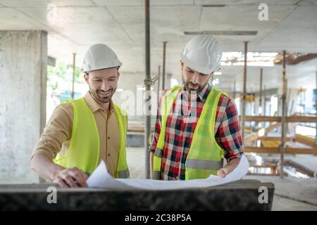Building site. Building workers in yellow vests brainstorming over blueprint, discussing it, smiling Stock Photo