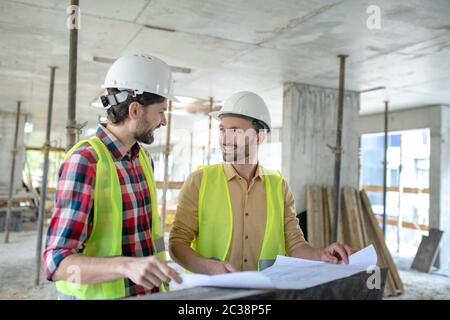 Building site. Building workers in yellow vests brainstorming over blueprint, discussing it Stock Photo
