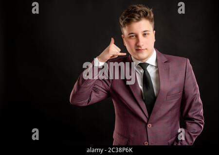 Law student with blond hair dressed in burgundy jacket, white shirt and black tie showing call me gesture posing on isolated black background with cop Stock Photo
