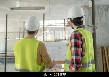 Building site. Building workers in yellow vests standing back to camera, holding blueprint, checking it Stock Photo
