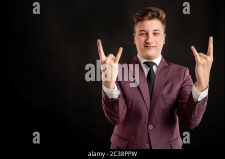 Law student with blond hair dressed in burgundy jacket, white shirt and black tie doing rock symbol with hands up posing on isolated black background Stock Photo