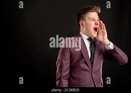 Law student with blond hair dressed in burgundy jacket, white shirt and black tie shouting with his hand to his mouth posing on isolated black backgro Stock Photo