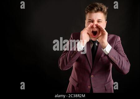 Law student with blond hair dressed in burgundy jacket, white shirt and black tie shouting with his hands to his mouth posing on isolated black backgr Stock Photo