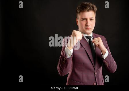 Law student with blond hair dressed in burgundy jacket, white shirt and black tie showing fists, boxing posing on isolated black background with copy Stock Photo
