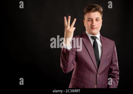 Law student with blond hair dressed in burgundy jacket, white shirt and black tie counting three with his finger posing on isolated black background w Stock Photo