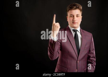 Law student with blond hair dressed in burgundy jacket, white shirt and black tie counting one with his finger posing on isolated black background wit Stock Photo