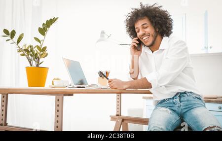 Side view of young entrepreneur or student talking mobile phone while working from home. Charming Arab guy chatting on smart phone with friends or Stock Photo