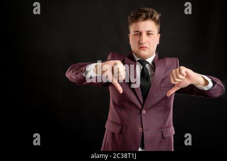 Law student with blond hair dressed in burgundy jacket, white shirt and black tie showing double dislike posing on isolated black background with copy Stock Photo