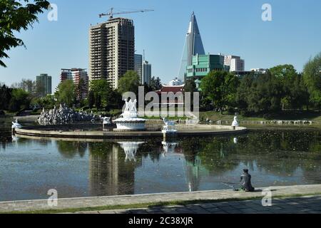 Pyongyang, North Korea - May 2, 2019: Pyongyang suburb. Water pond with fountain, park and local man fishing at morning time. Construction site and fa Stock Photo