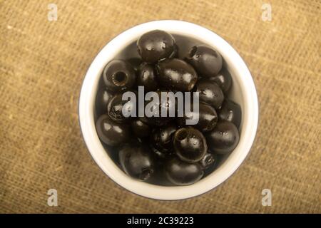 Black seedless olives in a ceramic bowl on a background of coarse-textured fabric. Close up Stock Photo