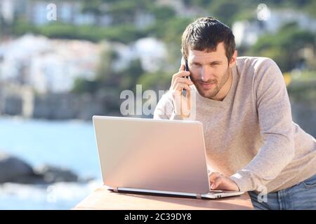 Serious man using a laptop and talking on mobile phone in a balcony on the beach Stock Photo