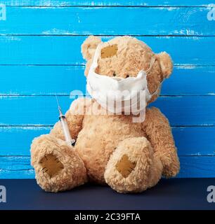 beige big teddy bear with patches sitting in a white medical mask in the paw of a syringe, concept of the need for vaccinations in children, blue wood Stock Photo