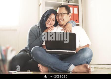 Muslim couple, smiling happily while looking at laptop, new married planning their life concept, sitting on floor of their new home Stock Photo