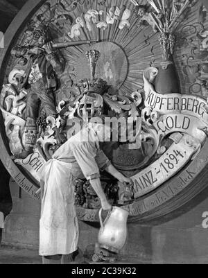 A winemaker with an apron taps wine from a decorated wine barrel. The following inscriptions are visible on the wine barrel: ' Kupferberg Gold', 'Built by Fr. Vlasdeck' and 'Mainz 1894'. In addition, a sun, a small angel, a man with a rifle and a crown are depicted on the front of the wine barrel. Stock Photo