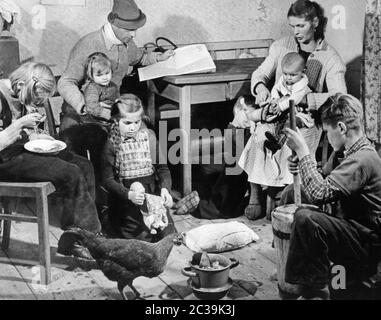 A family with five children in a slum in the Bavarian Forest. A chicken runs through the room. One of the girls is eating something, another one is playing with a doll and the boy is churning milk. Stock Photo