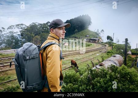 Portrait of young man with backpack against train station in clouds. Idalgashinna, Sri Lanka. Stock Photo