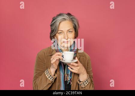 Charming Caucasian woman drinks coffee holding white cup. Pretty mature lady smiles slightly isolated on pink background. Beauty concept. Coffee time Stock Photo