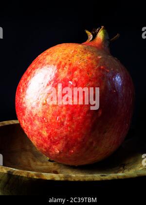 A single washed pomegranate with water droplets in a wooden dish against a black background