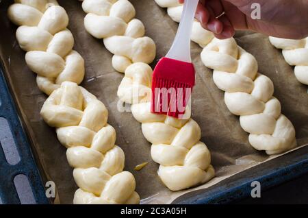 Preparation process - raw unbaked buns. Female hand lubricates raw buns with an egg before baking. Rustic style. Traditional pastry Stock Photo