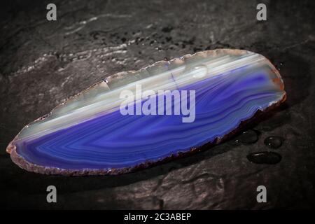 Close-up of an elongated flat polished agate slice with blue and white stripes lying on a slate plate with water drops. Stock Photo