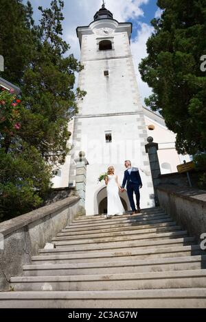 The Kiss. Bride and groom holding hands walking down the staircase in front of a small local church. Stylish wedding couple kissing. Stock Photo