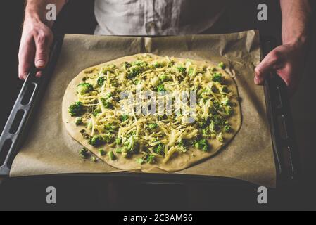 Man holding baking dish with uncooked pizza with broccoli, pesto sauce, spices and cheese Stock Photo