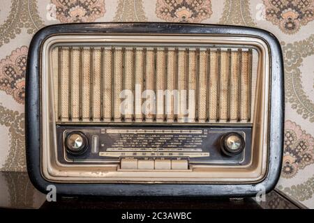 An old transistor radio, with knobs and buttons for manual tuning. In the background a vintage wallpaper. Ancient object, worn and ruined by time. Stock Photo