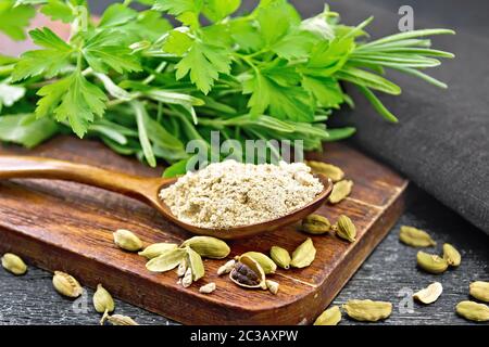 Ground cardamom in a spoon, seasoning capsules, napkin, fresh parsley and rosemary on black wooden board background Stock Photo