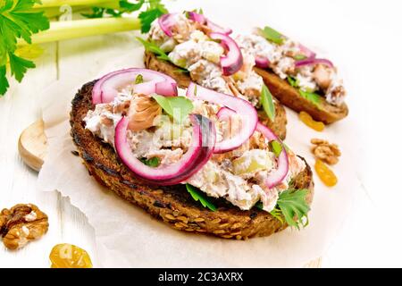 Salmon, petiole celery, raisins, walnuts, red onions and curd cheese salad on toasted bread with green lettuce leaves on parchment on a light wooden b Stock Photo