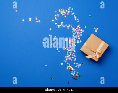 closed square golden gift box on a blue background, near multi-colored sequins, top view, festive backdrop Stock Photo
