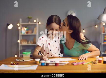 The child draws a drawing. Mother teaches daughter to draw with paints on paper sitting at a table in a living room. Stock Photo