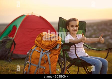 Cute little girl showing thumbs up gesture during camping trip in mountains. Child enjoying summer vacation Stock Photo