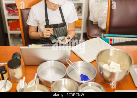 Female baker using cell phone with laptop and bakery ingredient on table. Stock Photo