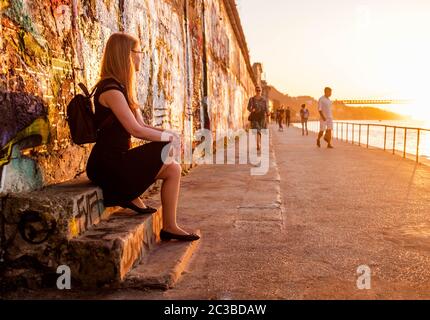 Tourist girl with a backpack sitting on dilapidated stairs of an old, abandoned factory and people watching with sunset scenery on a background. Stock Photo