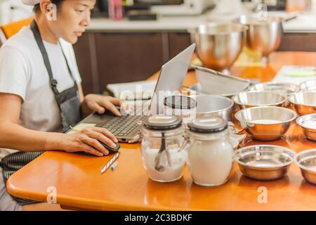 Female baker working on laptop with bakery ingredient on table. Stock Photo